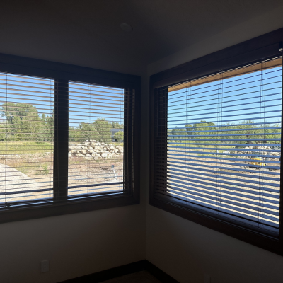 A corner of a room with two large windows framed in dark wood, covered by horizontal blinds. The blinds are partially open, revealing a bright outdoor scene with a rock pile, green trees, and construction equipment under a clear blue sky.